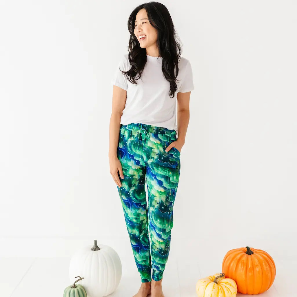 Smiling woman in a white T-shirt wearing green and blue wave-patterned pants standing barefoot next to pumpkins on a white background.