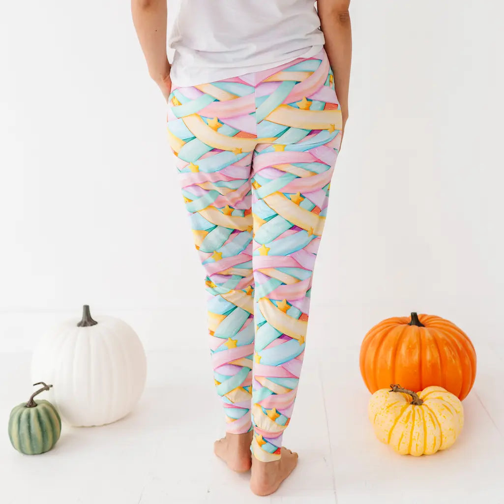 Back view of a person wearing pastel candy ribbon-patterned pants and a white shirt standing barefoot among pumpkins on a white background.