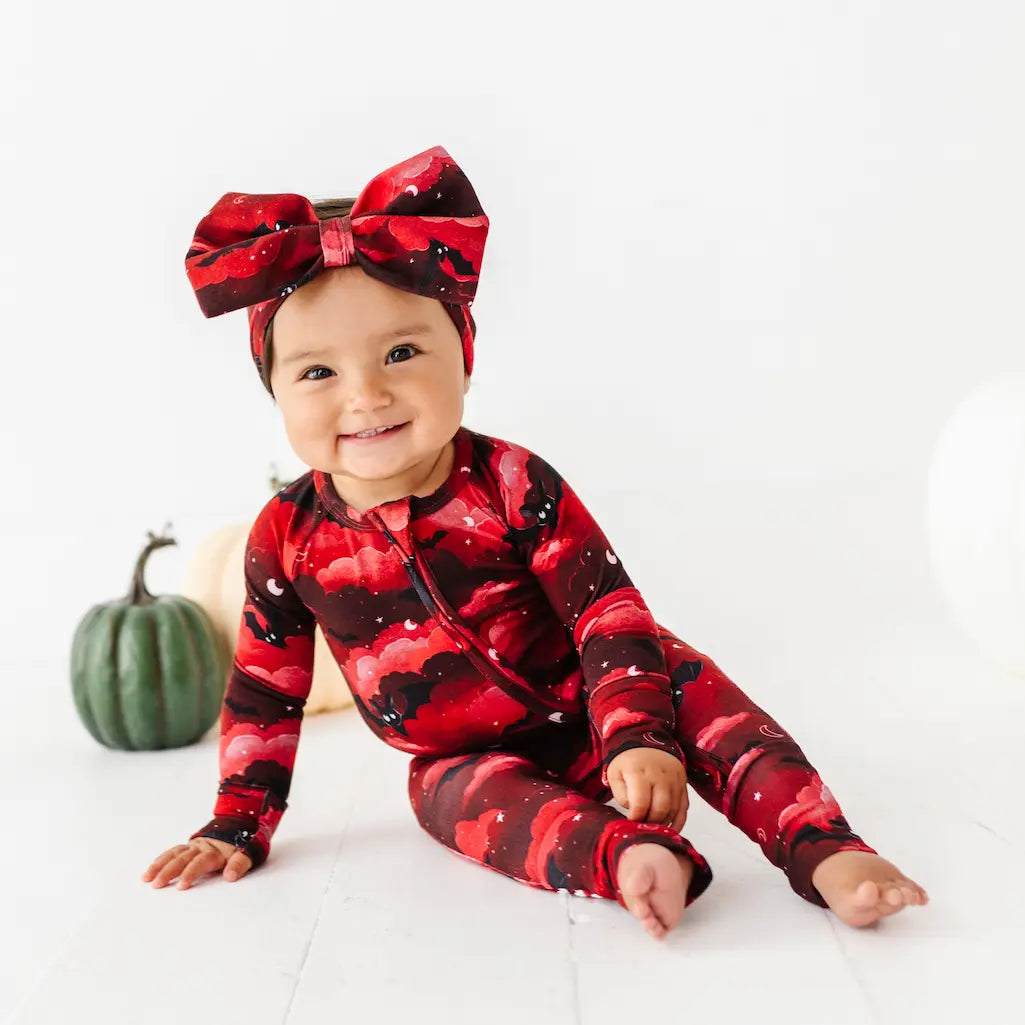 Baby wearing a red and black patterned onesie with a matching headband, sitting on a white surface with a small green pumpkin.