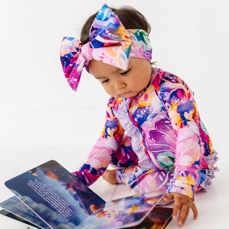 Child wearing a colorful outfit and headband, sitting with a book on a white background