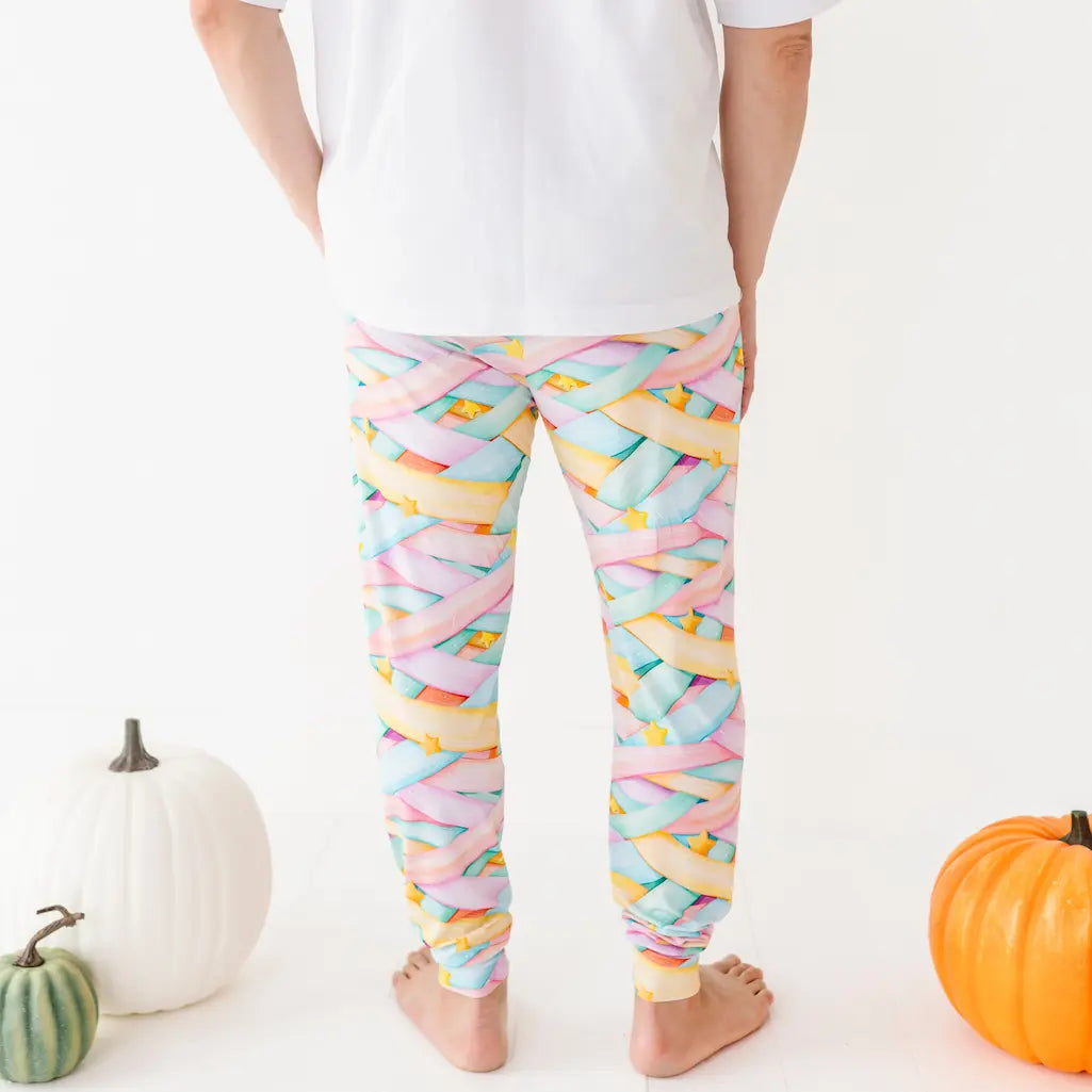 Back view of a person wearing colorful pastel candy ribbon-patterned jogger pants and a white shirt standing barefoot among pumpkins on a white background.