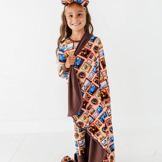 A smiling child in a bow headband holding a blanket with a framed classic paintings print and brown reverse side.