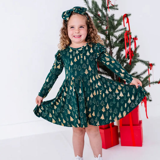 A child with a big smile holding out the skirt of a long sleeve twirl dress with flutter ruffles at the shoulders in a dark green print of gold Christmas trees.