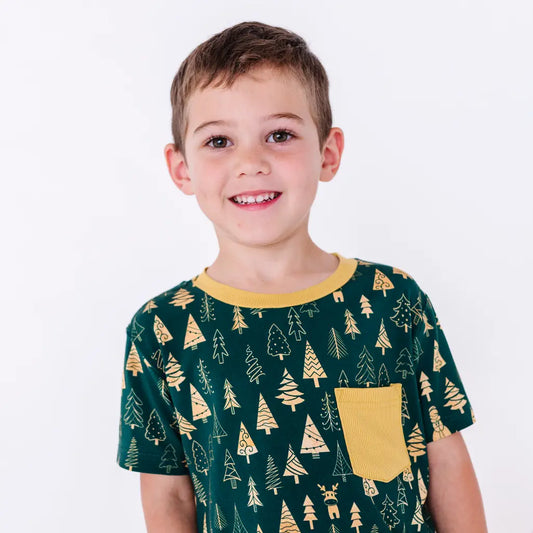 A child with a big smile looking at the camera, wearing a short sleeve pocket tee with a mustard ribbed collar and pocket in a dark green print of gold Christmas trees.