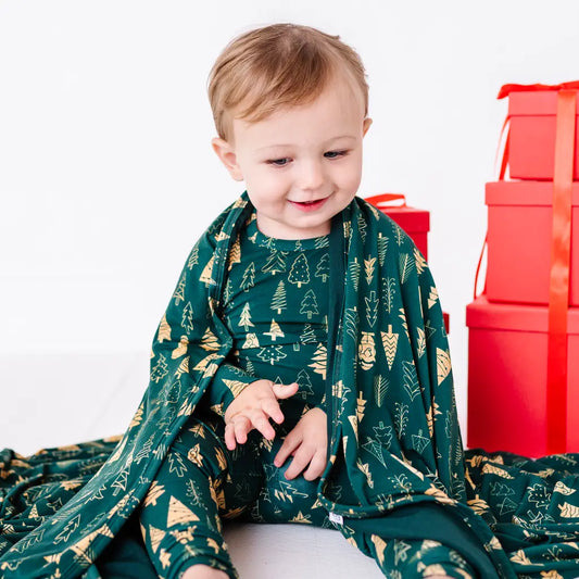 A smiling toddler in matching pajamas sitting on the floor with a dark green blanket in a print of gold and outlined Christmas trees draped over the shoulders and spread around.