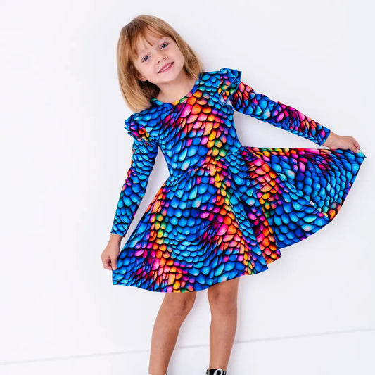 A smiling child holding out the skirt of a long sleeve twirl dress with flutter ruffles at the shoulders in a rainbow dragon scale print of blue, pink, orange, and yellow.