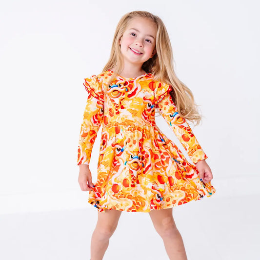 A child with a big smile holding out the skirt of a long sleeve twirl dress with flutter ruffles at the shoulders, in a golden yellow print of lion dance heads and red lanterns.