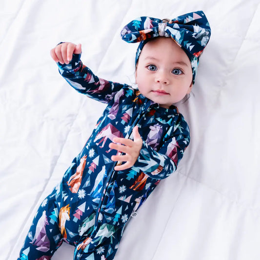 A baby lying on a white quilt with one arm raised, wearing a large fabric bow headband in a navy geometric wolf print with triangle pine trees and snowflakes, with a matching zip up pajama.