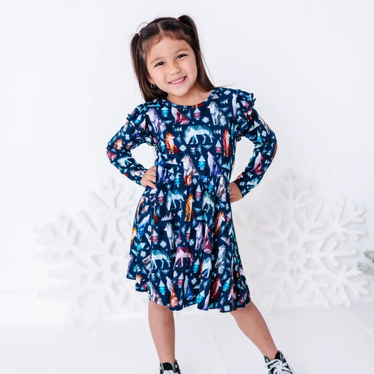 A child smiling with hands on the hips in a long sleeve twirl dress with flutter ruffles at the shoulders in a navy print of geometric wolves, triangle pine trees, and snowflakes.