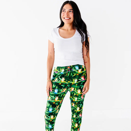 A woman in a white tee smiling with her hand in the pocket of dark green pajama pants with a colorful tree frog print.