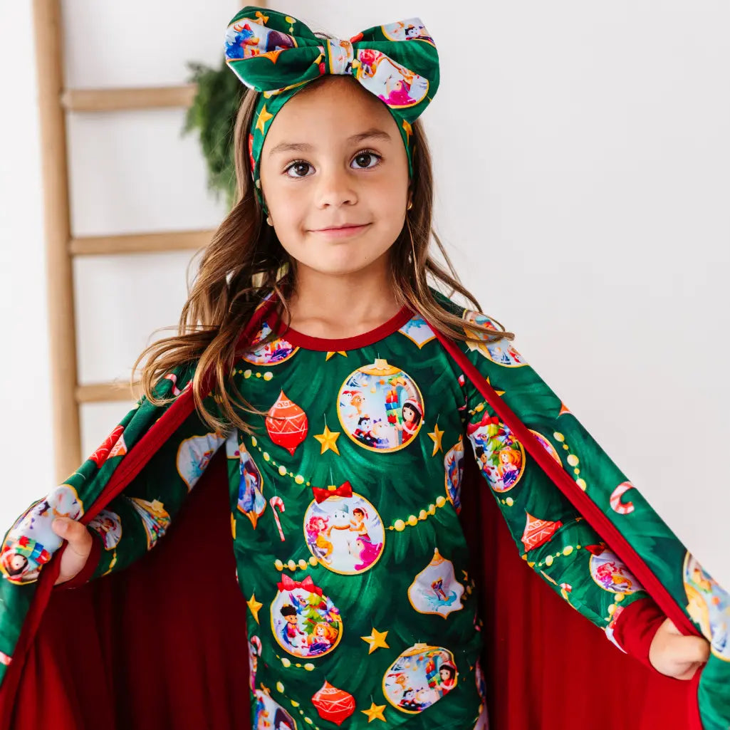 Child wearing a festive green pajama set with a red cape and headband, standing against a white background.