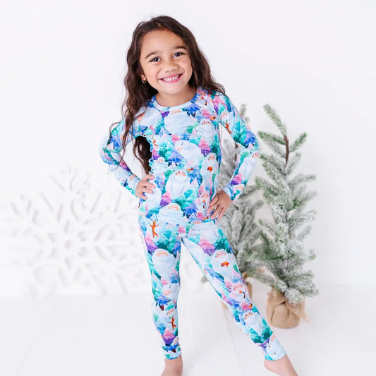 A smiling child with hands on hips in a long sleeve pajama set with a print of friendly white yetis among teal, blue, and purple pine trees, standing beside small frosted trees.