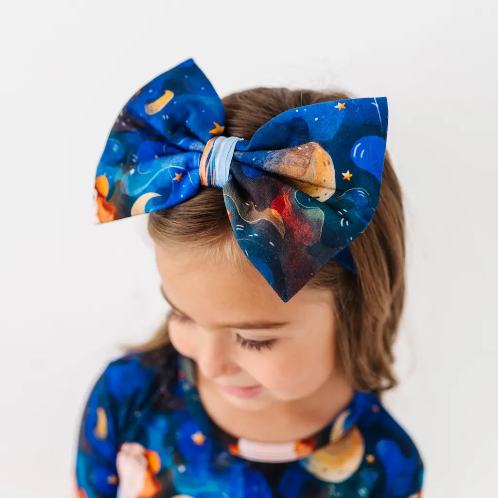 Child wearing a large navy blue bow headband featuring a colorful space-themed pattern with planets and stars and a matching outfit, looking down against a white background.