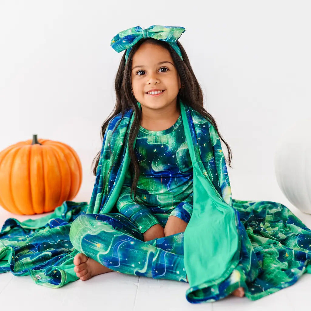 Young girl in a green and blue patterned outfit with matching headband, sitting on the floor with pumpkins beside her.
