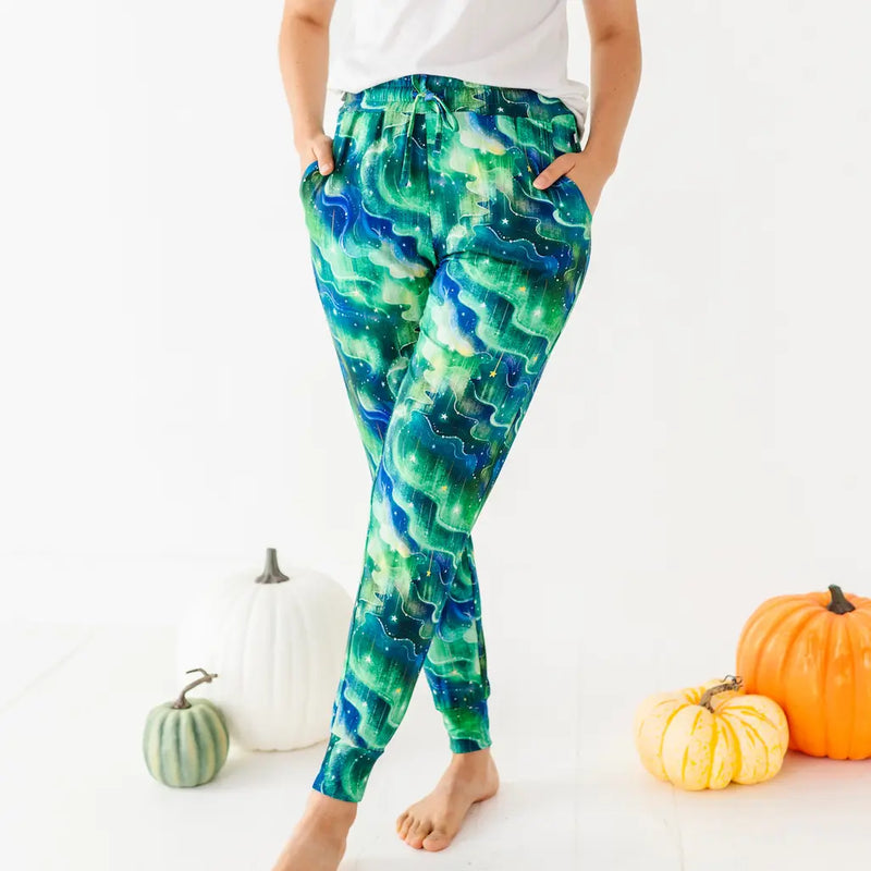 Person wearing green and blue wave-patterned pants with hands in pockets and crossed ankles standing by pumpkins on a white background.