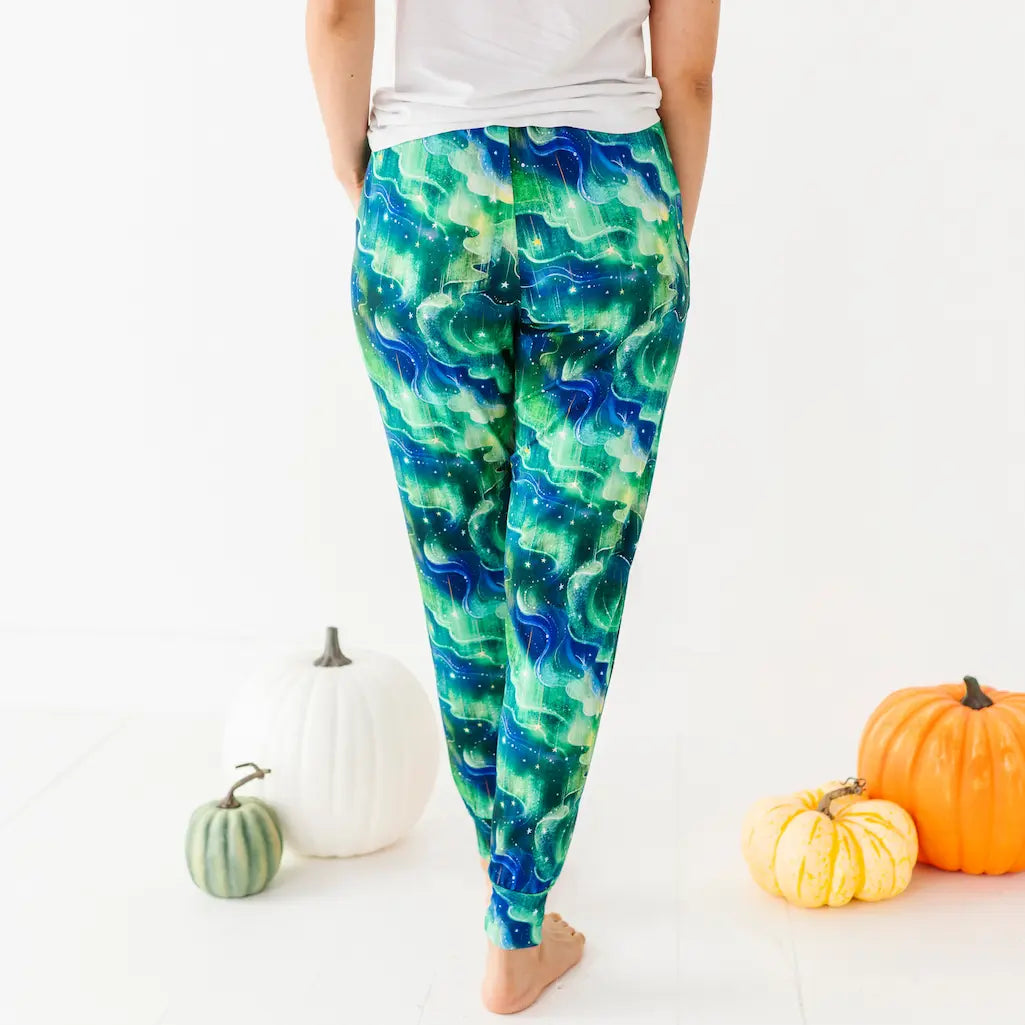 Rear view of a person wearing green and blue wave-patterned leggings and a white shirt standing barefoot among pumpkins on a white background.