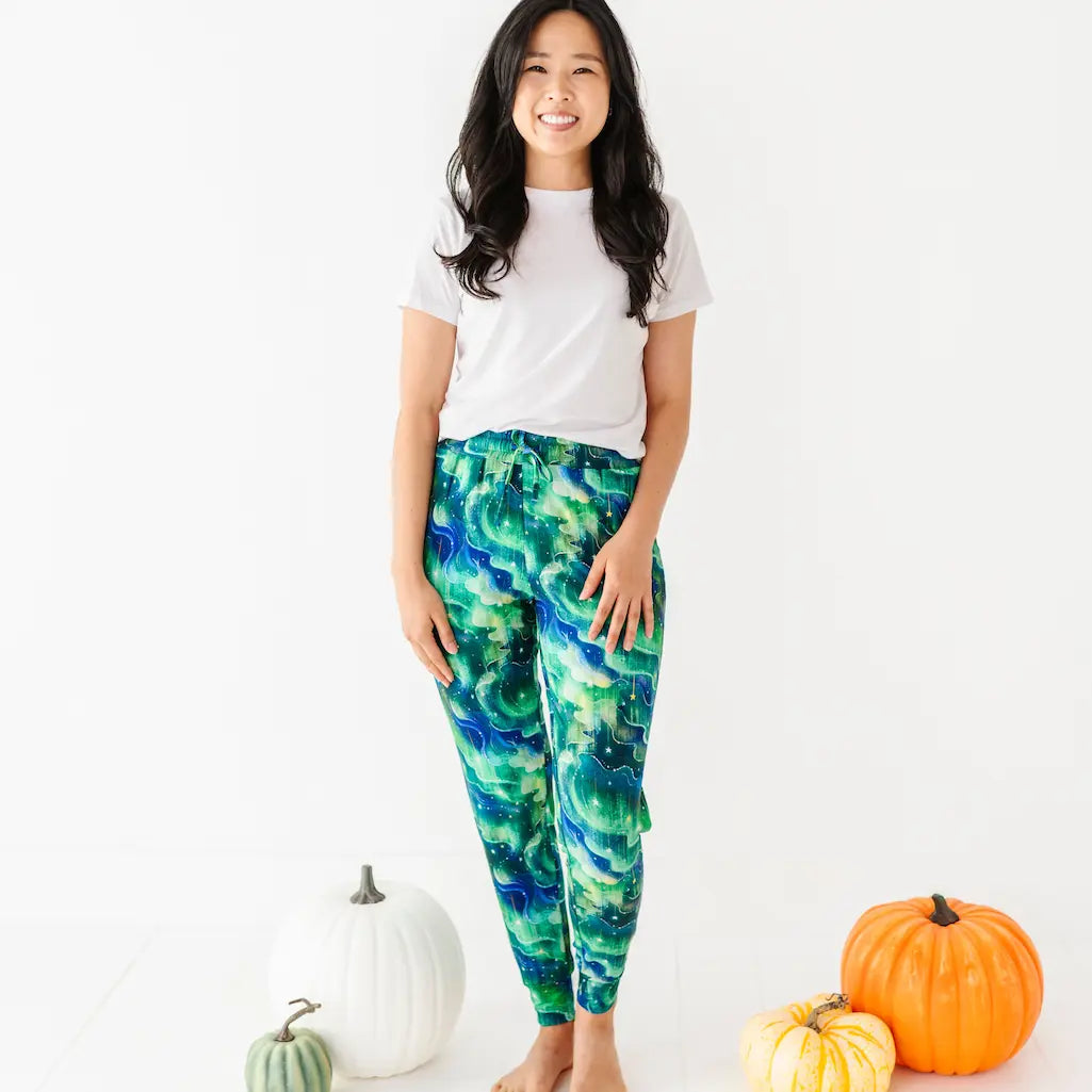 Woman in a white T-shirt and green-blue wave patterned pants standing barefoot next to pumpkins on a white background.