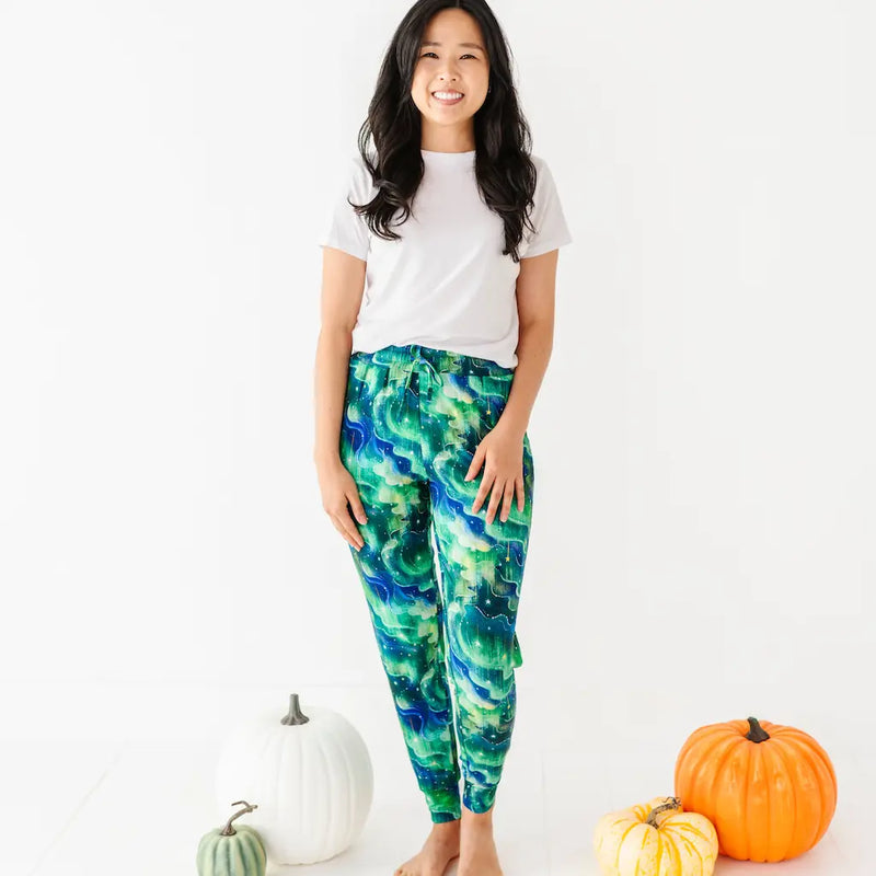 Woman in a white T-shirt and green-blue wave patterned pants standing barefoot next to pumpkins on a white background.