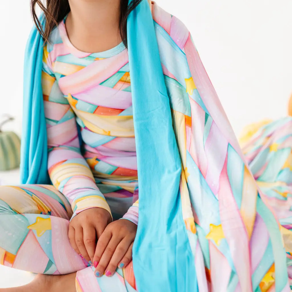 Close-up of a child wearing pastel candy ribbon-patterned long-sleeve pajamas with a turquoise blanket draped over one shoulder, seated cross-legged on a white surface with small pumpkins in the background.