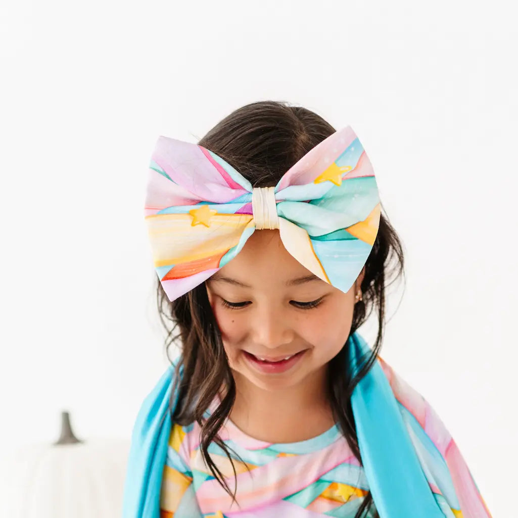 Child wearing a colorful oversized pastel headband bow with yellow stars and a matching pastel-patterned outfit, looking down against a white background.