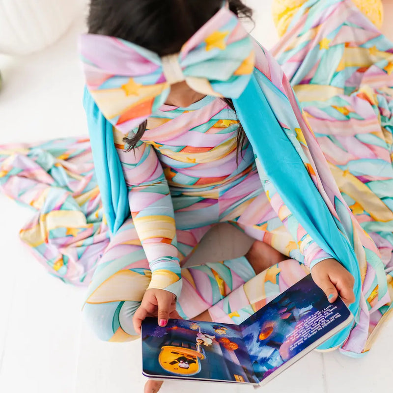 Child wearing a colorful pastel candy ribbon-patterned long-sleeve outfit with a large matching bow headband and turquoise cape, sitting on a white surface while reading an illustrated Halloween book.
