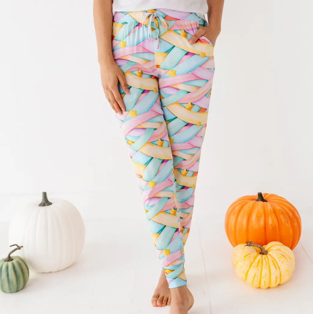 Person wearing colorful pastel candy ribbon-patterned leggings with a drawstring and yellow stars, hands in pockets, standing barefoot among pumpkins on a white background.
