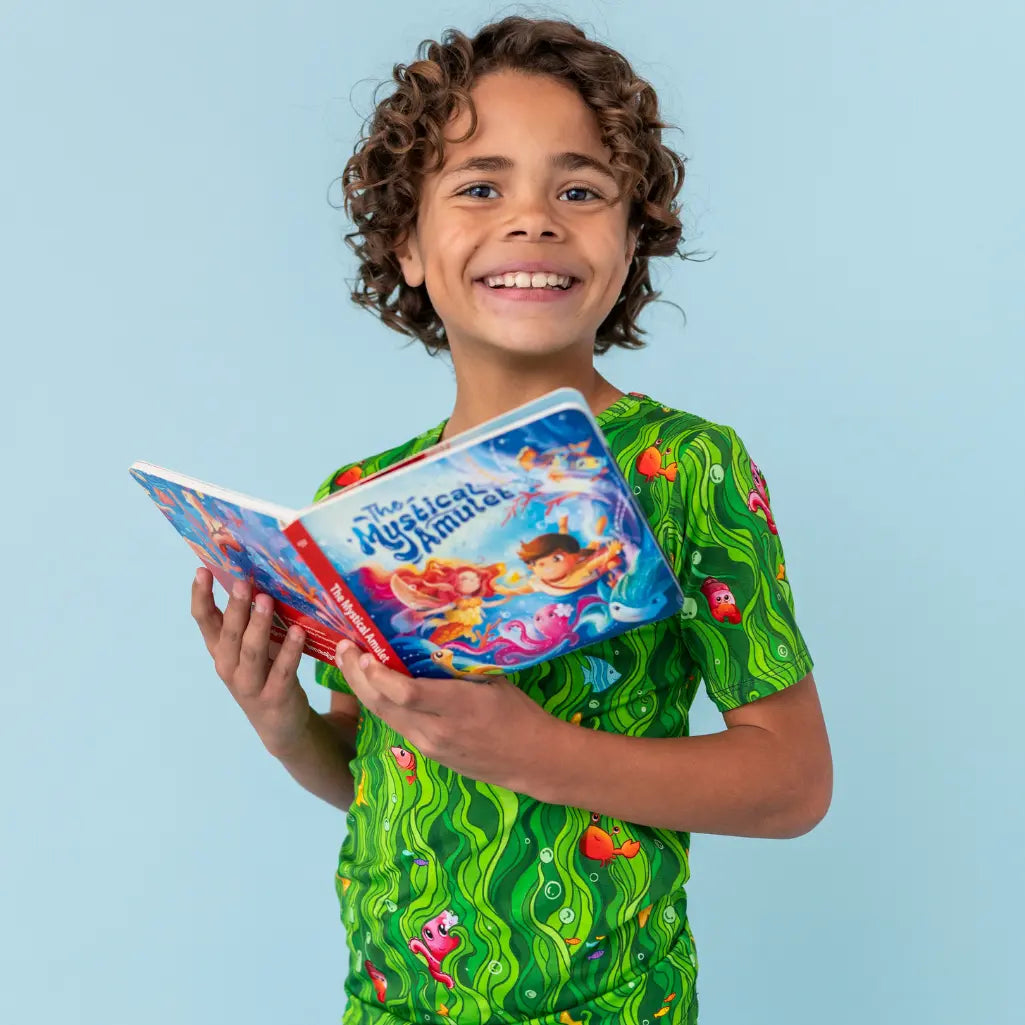 Child wearing a green shirt with cartoon designs, holding a book titled 'The Magical Mermaid', against a light blue background.