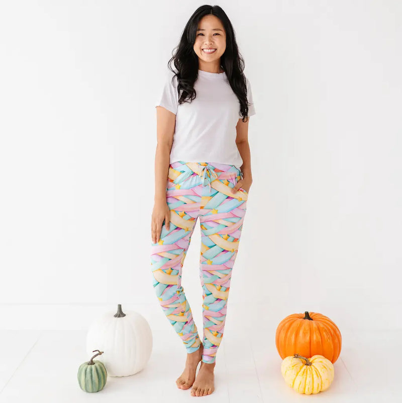 Woman in a white T-shirt wearing colorful pastel candy ribbon-patterned leggings with a drawstring and yellow stars, standing barefoot among pumpkins on a white background.