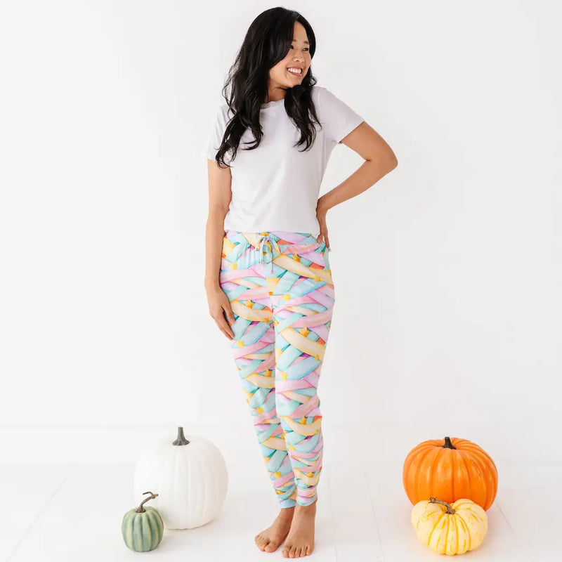 Smiling woman in a white T-shirt wearing colorful pastel candy ribbon-patterned joggers with a drawstring, standing barefoot with one hand on her hip next to pumpkins on a white background.