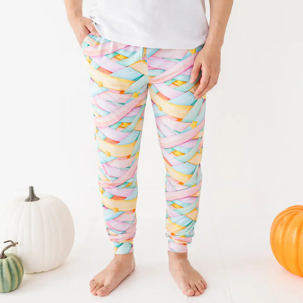 Child wearing pastel candy ribbon-patterned leggings standing barefoot with hands in pockets next to pumpkins on a white background.