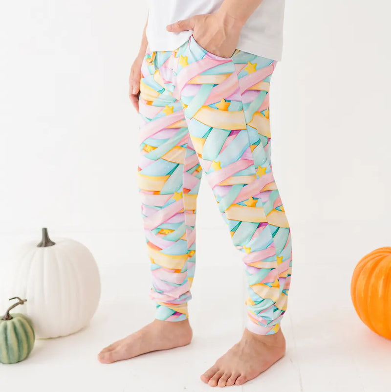 Person wearing colorful pastel candy ribbon-patterned leggings standing barefoot with hands in pockets next to pumpkins on a white background.
