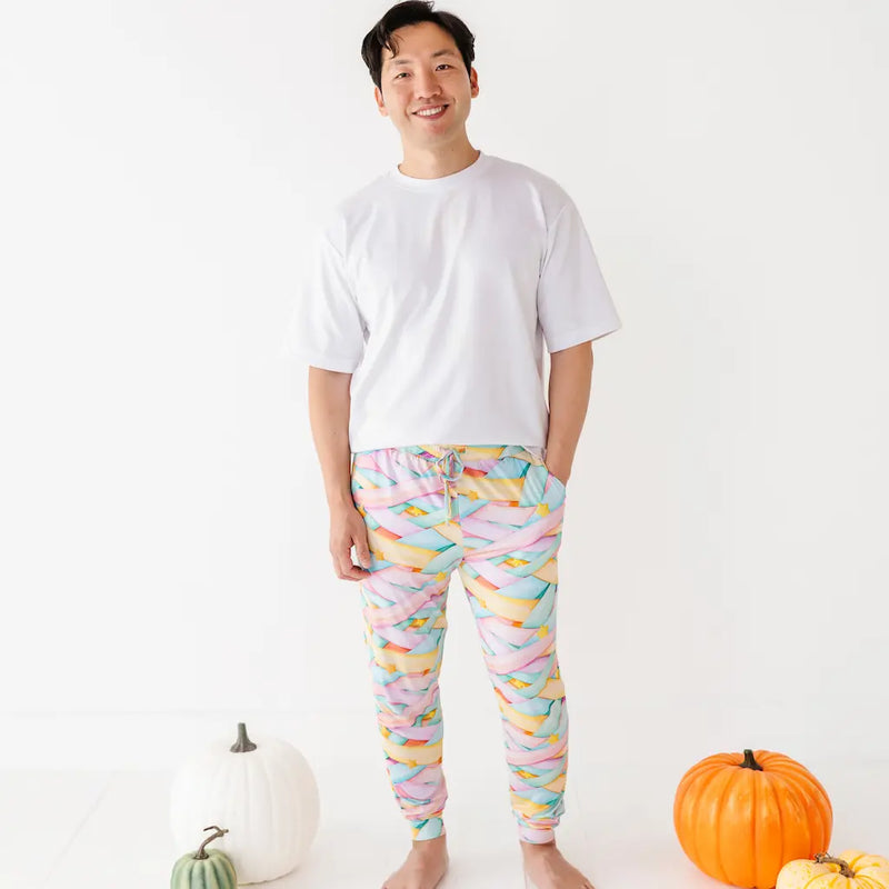 Smiling man in a white T-shirt wearing colorful pastel candy ribbon-patterned pajama pants with a drawstring and yellow stars, standing barefoot next to pumpkins on a white background.