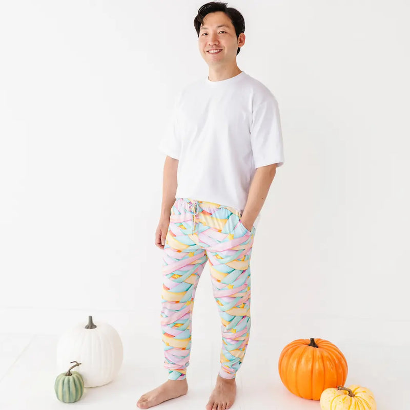 Smiling man in a white T-shirt wearing colorful pastel candy ribbon-patterned jogger pants with a drawstring, hands in pockets, standing barefoot among pumpkins on a white background.
