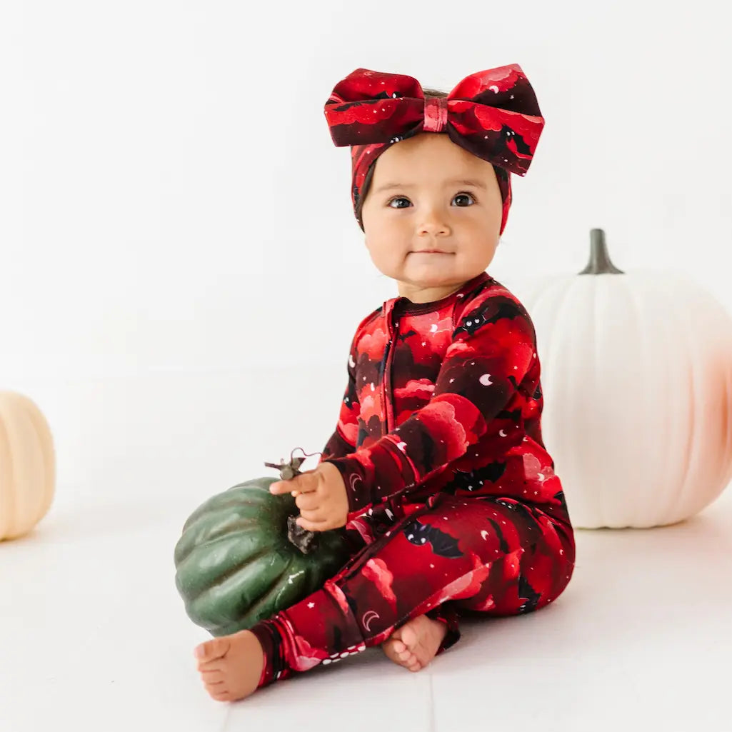 Baby in red and black patterned outfit with a large bow, holding a small pumpkin.