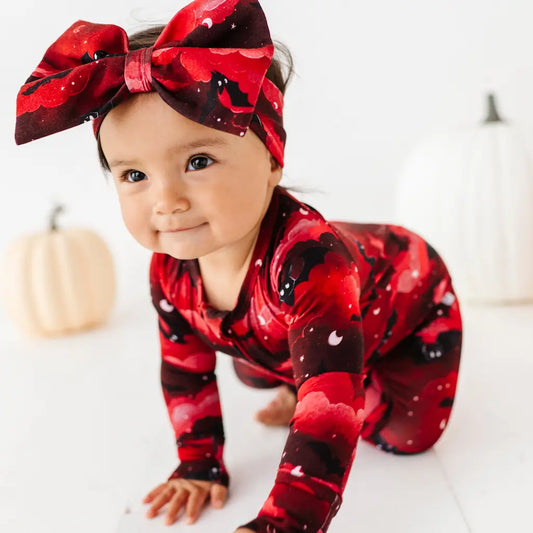 Baby in red and black patterned outfit with a large bow, standing on a white background.