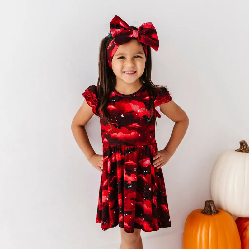 Young girl wearing a red dress with black patterns and a matching headband, standing next to pumpkins.