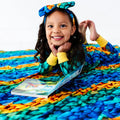 Child lying on a colorful blanket with a book, wearing a large bow headband.