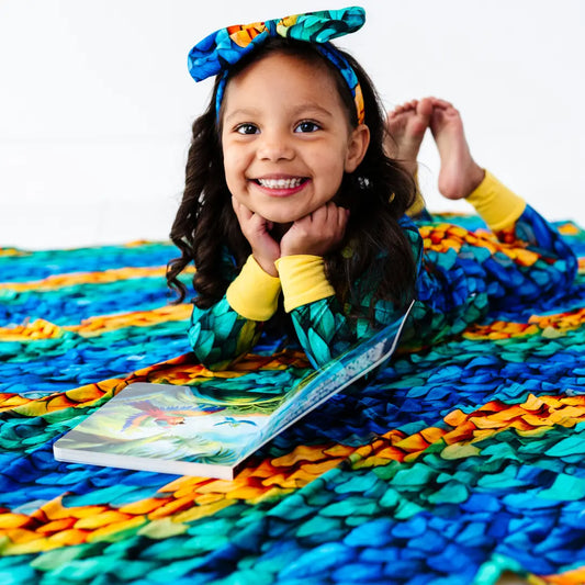 Child lying on a colorful blanket with a book, wearing a large bow headband.
