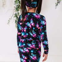 Back view of a child in a black pajama set with glowing pink and blue butterflies and a matching headband.