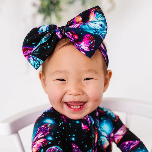 A smiling baby wearing a multicolored bow headband with a pattern of twinkling stars and shimmering wings.