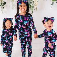 Three children holding hands in matching black pajamas with glowing pink and blue butterflies and bow headbands.