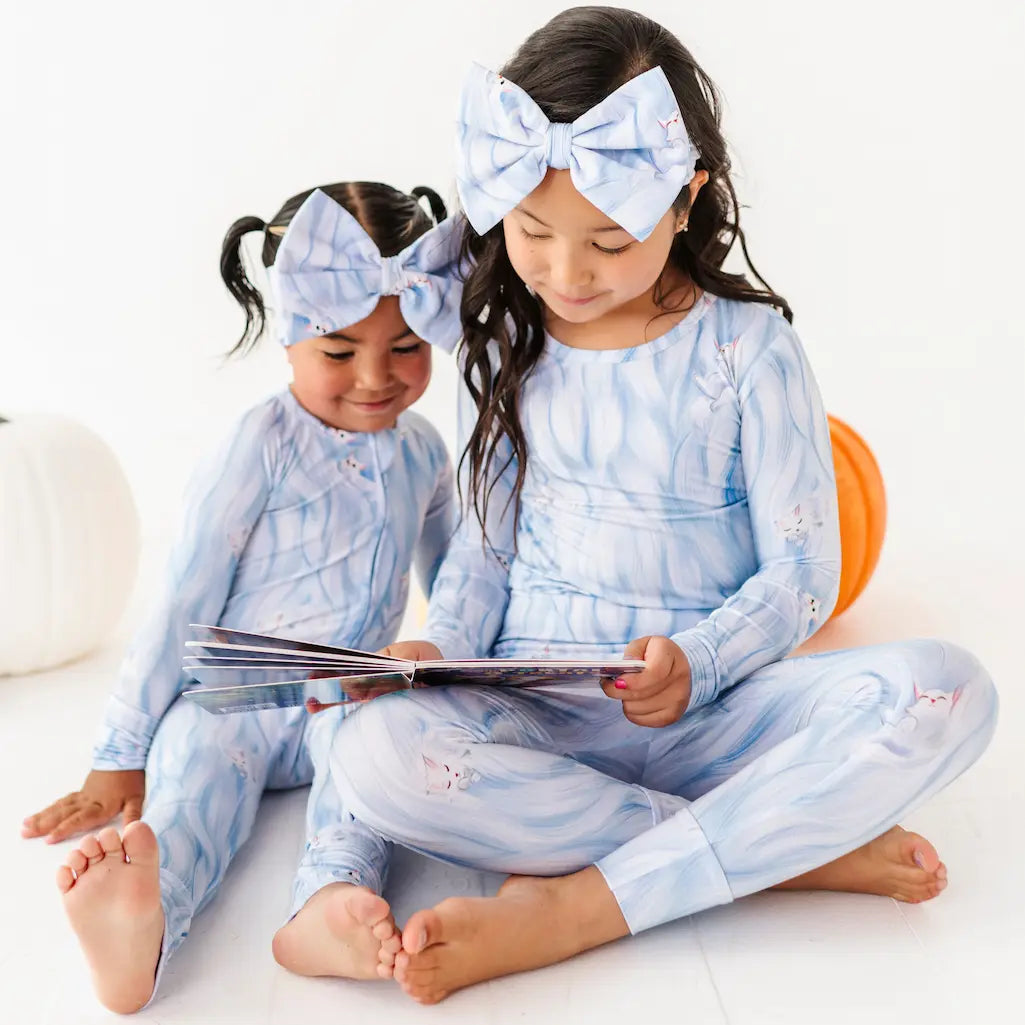 Two children in matching blue pajamas with large bows sitting on a white surface.