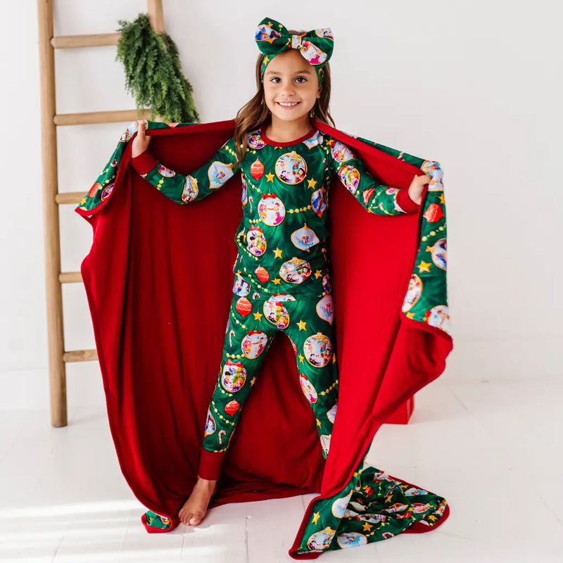 Child wearing a festive green pajama set with a red blanket, standing in front of a white wall.