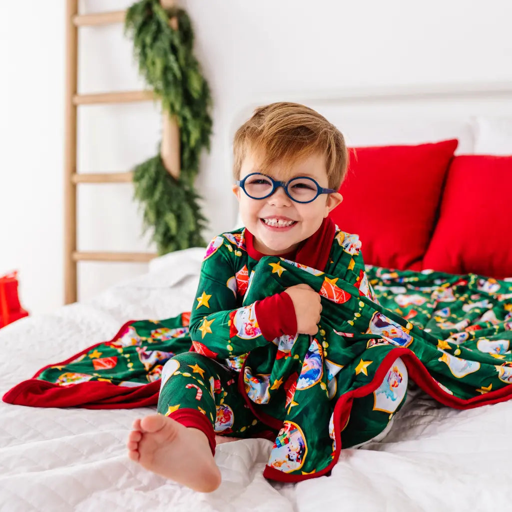 Child wearing a festive green pajama set with red accents, sitting on a bed with a Christmas tree in the background.