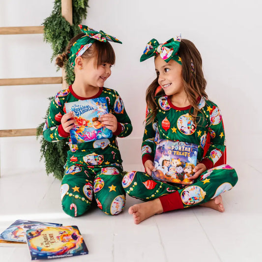 Two children in festive pajamas with holiday-themed books, sitting on a white floor.