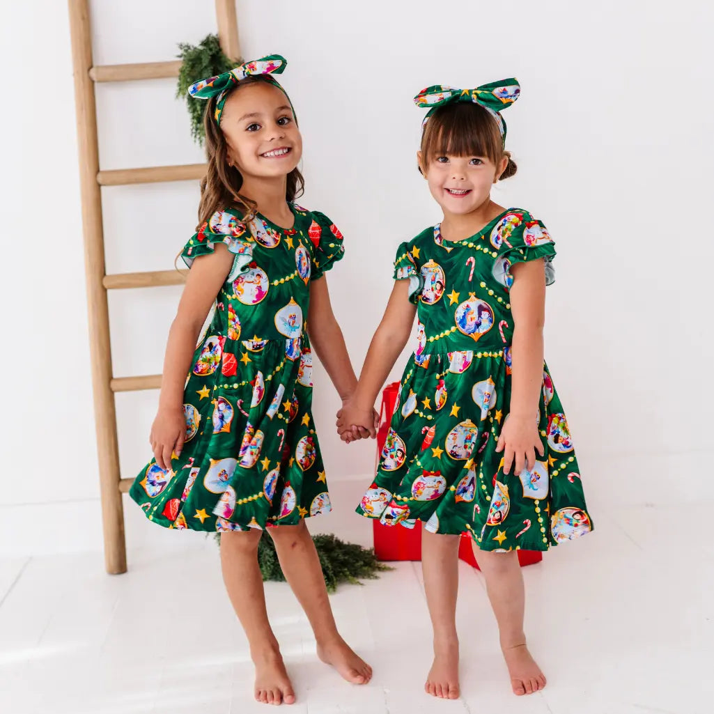 Two young girls wearing green dresses with festive patterns, holding hands against a white background.