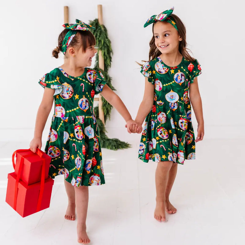 Two young girls wearing matching green dresses with festive patterns, holding hands against a white background.