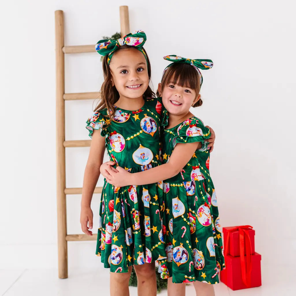Two children wearing matching green dresses with festive patterns, standing in front of a white wall with a wooden ladder and red gift bag.