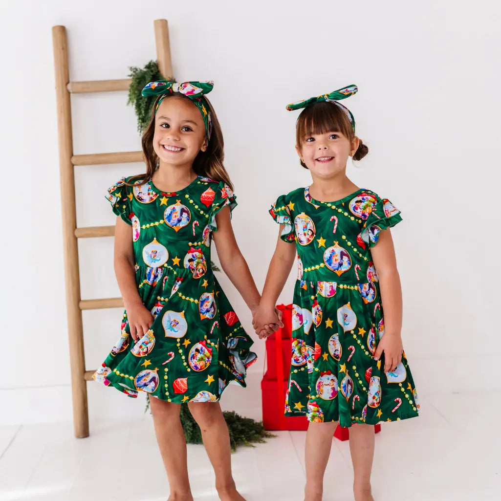 Two young girls wearing matching green dresses with festive designs, holding hands against a white background.