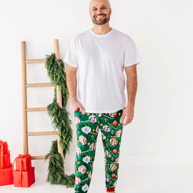 Man wearing a white t-shirt and green festive pajama pants with a ladder and Christmas decorations in the background.